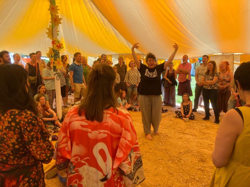 A woman stands in the centre of a circle of people holding her hands in the air conducting them in song, underneath the yellow and white stripes of a marquee
